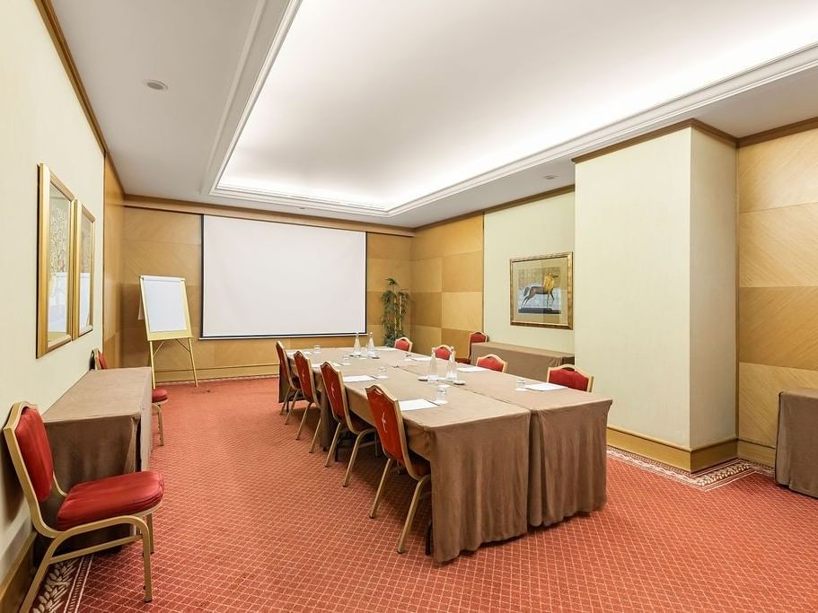 Sala de reuniones con mesa larga, sillas rojas, pizarra, proyector y pinturas en las paredes.