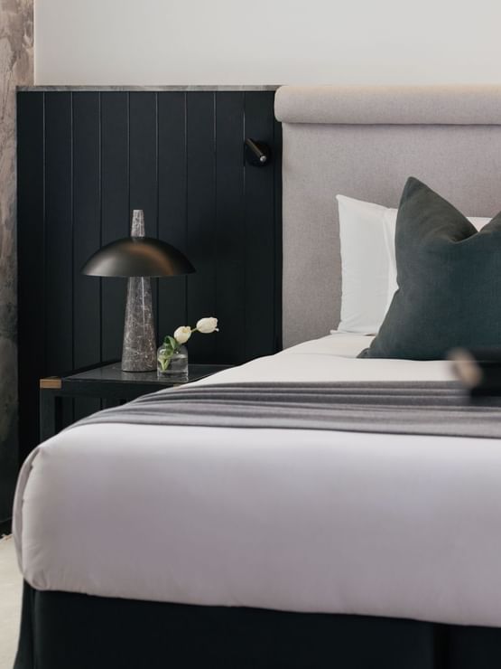 Elysium Suite at Elysium Manly with a black lamp and tulip vase by a wooden nightstand under a grey headboard