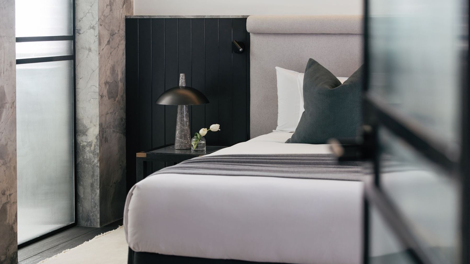 Elysium Suite at Elysium Manly with a black lamp and tulip vase by a wooden nightstand under a grey headboard