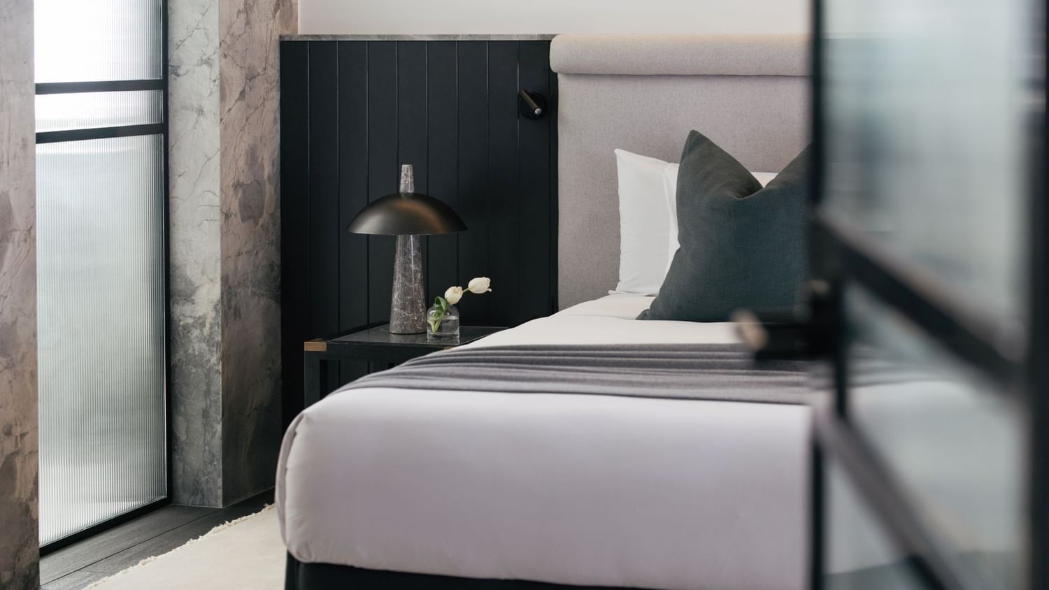 Elysium Suite at Elysium Manly with a black lamp and tulip vase by a wooden nightstand under a grey headboard