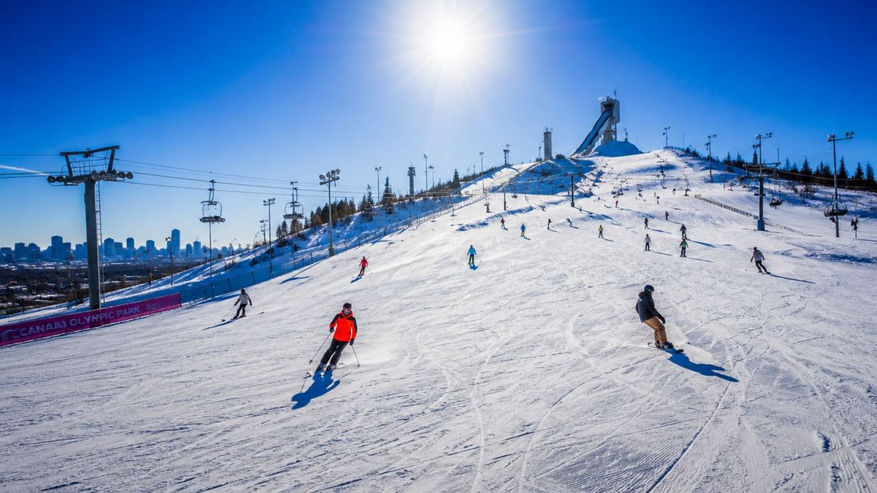 Skiers descending snowy slopes with chairlifts and a ski jump tower visible at Canada Olympic Park under a clear blue sky.