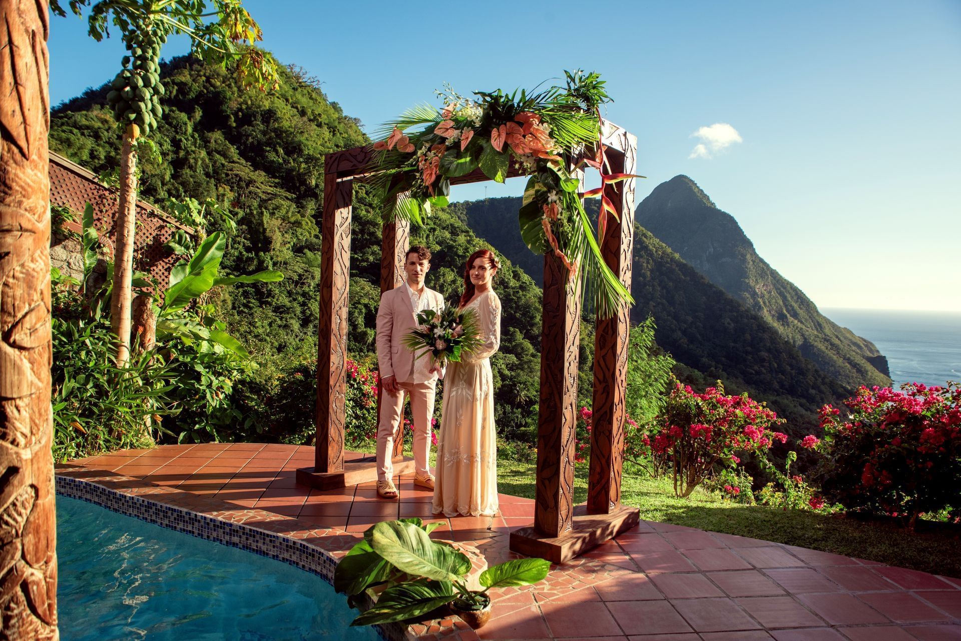 Couple standing under a floral arch near a pool with mountains in the background.