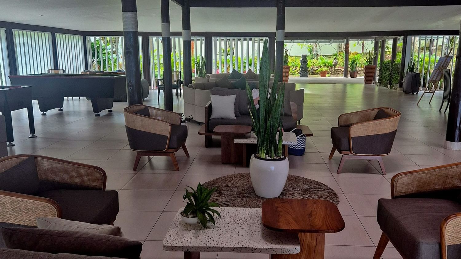 Spacious open-air lobby with modern seating, exposed beam ceiling, potted plants, and views of tropical greenery beyond.