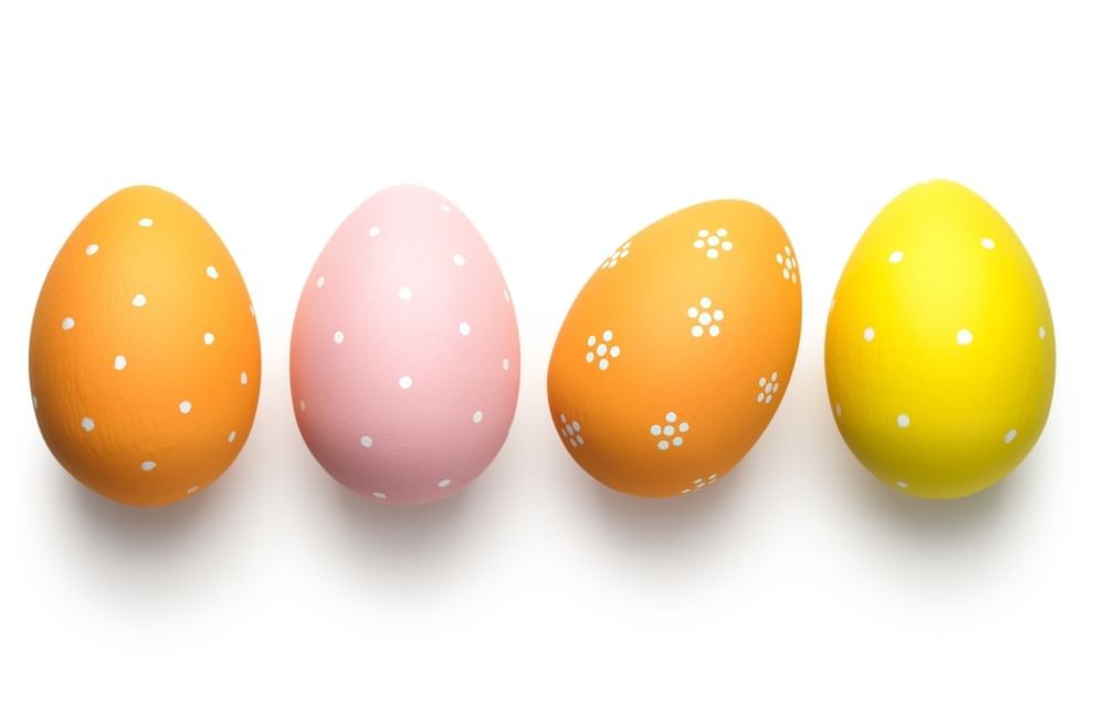 Four Easter Eggs, two orange, one pink, and one yellow, with small white polka dots and flowers, sit against a white background. 