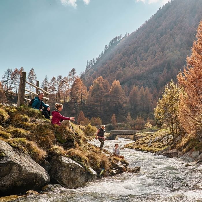 A family enjoying a sunny day by a river with mountain views and autumnal foliage.