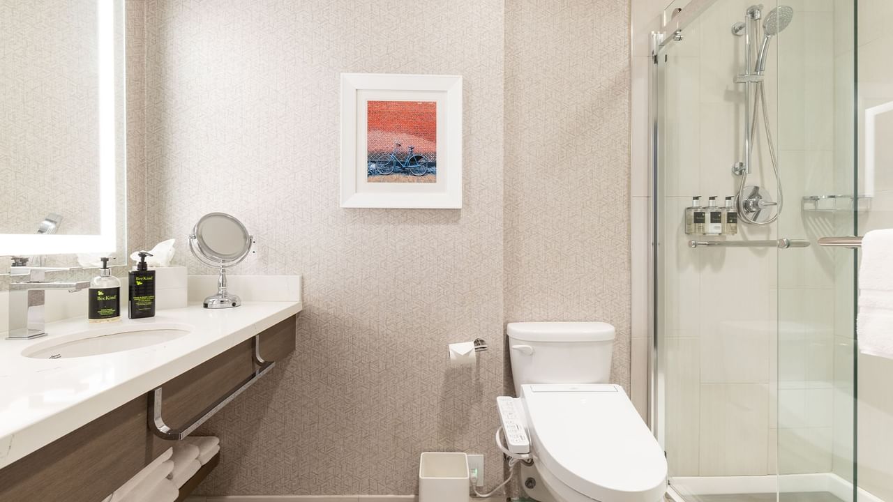 Hotel bathroom with toilet, sink, shower, framed picture, mirror, and folded towels on a shelf.