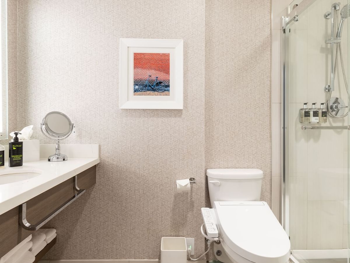 A modern hotel bathroom with a toilet, shower stall, sink, framed artwork, and toiletries.