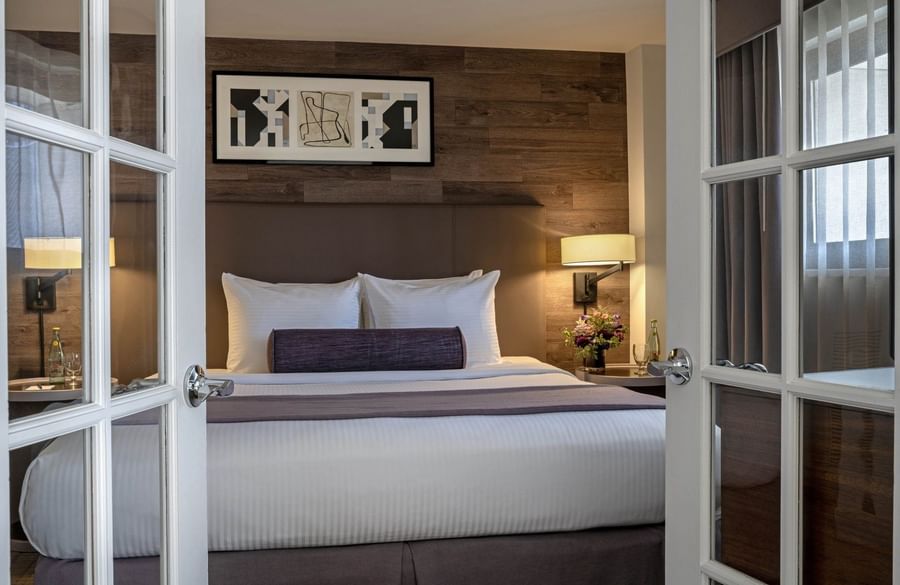 View through white french doors of a bed with a bolster by a bedside lamp in Premier Suite at Warwick Denver