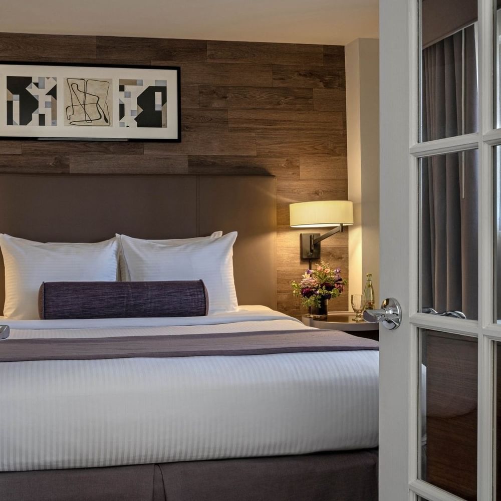 Plush bed with a purple bolster by a wall lamp seen through white French doors in Premier Suite at Warwick Denver