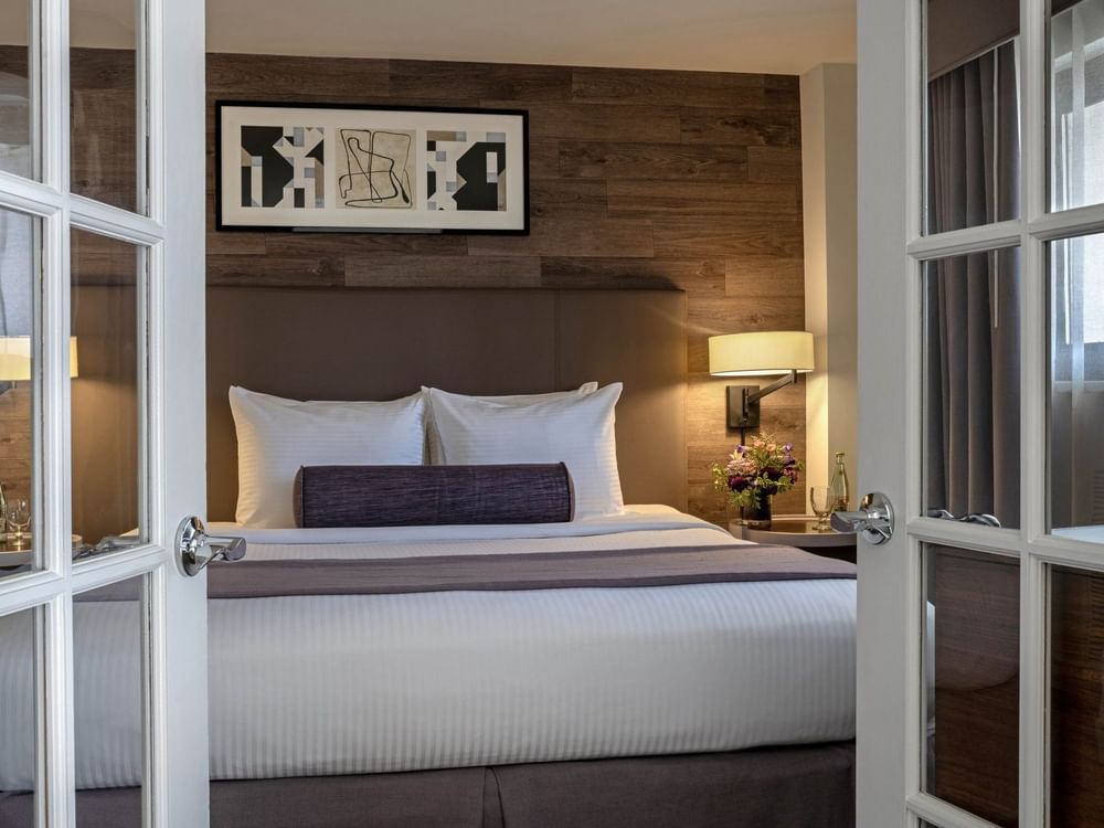 Plush bed with a purple bolster by a wall lamp seen through white French doors in Premier Suite at Warwick Denver
