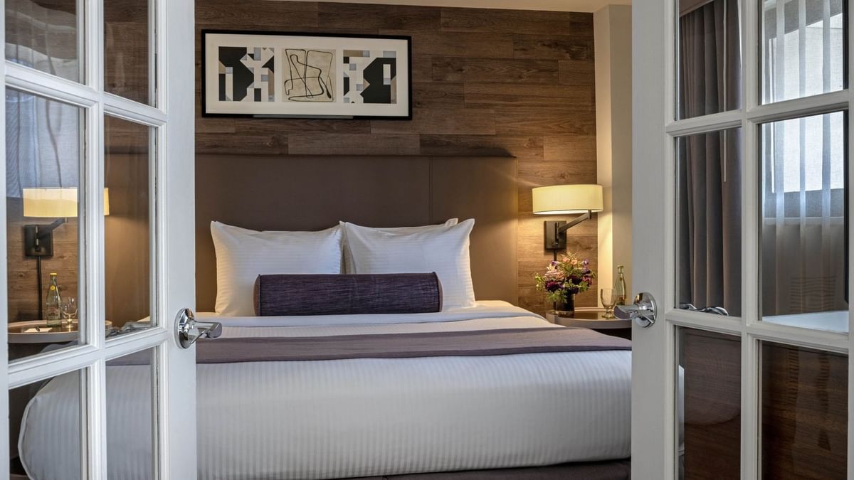 Plush bed with a purple bolster by a wall lamp seen through white French doors in Premier Suite at Warwick Denver