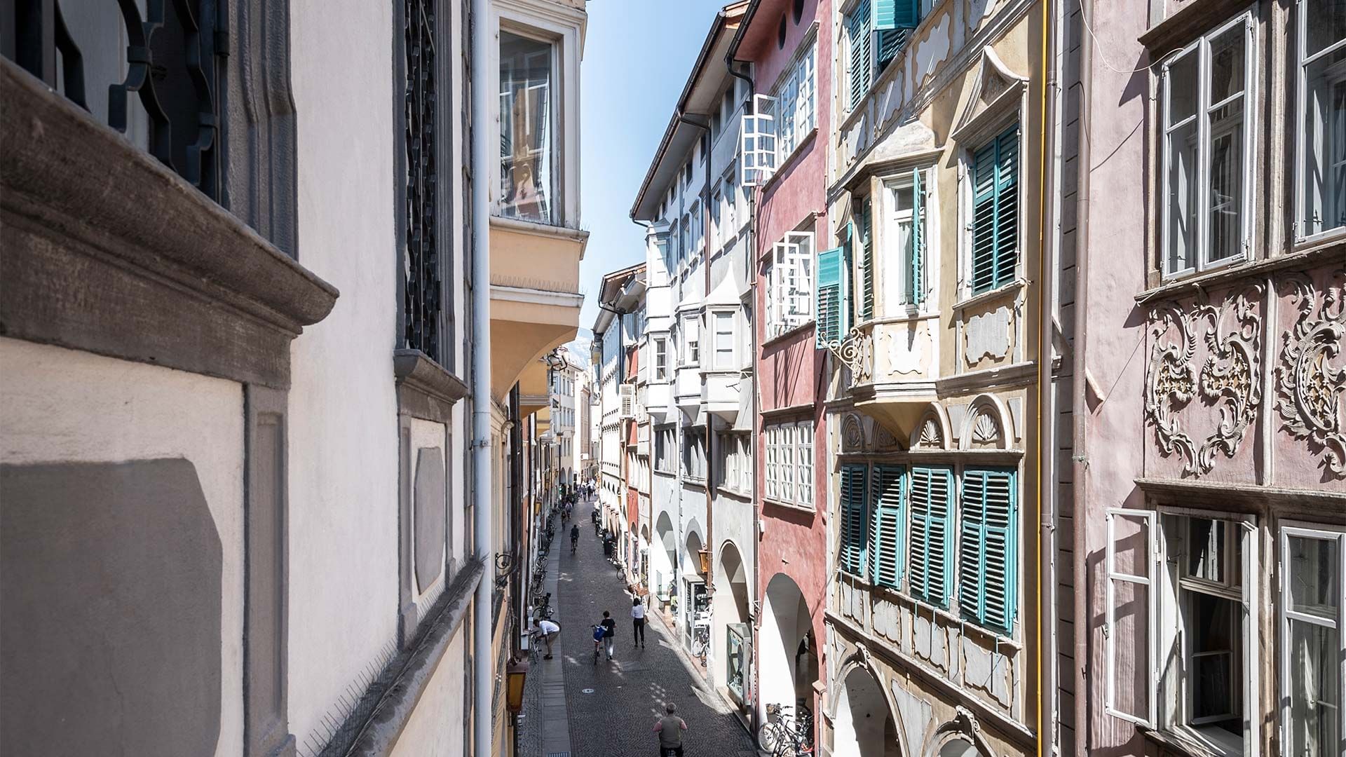 Strada stretta fiancheggiata da edifici decorati e persone che camminano sotto il sole.