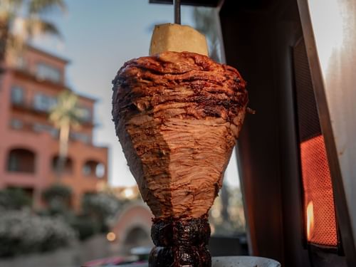 Spit-grilled meat for tacos al pastor cooking outdoors in front of a resort building in Tahona at Hacienda del Mar Los Cabos.