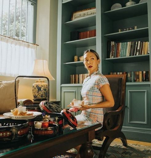 A woman sits at a table with snacks and tea surrounded by charming decor and a bookshelf at The Majestic Malacca