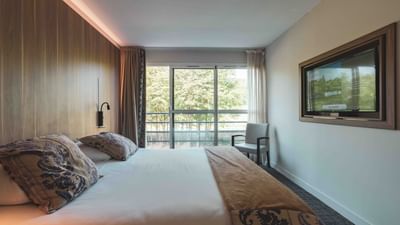 Modern hotel room with large bed, chair, TV, and balcony overlooking trees.