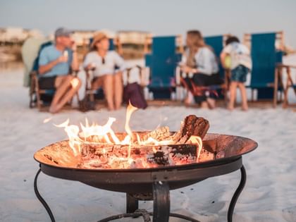 Beachside fire pit with flames burning while guests gather on chairs with blue privacy screens in the background