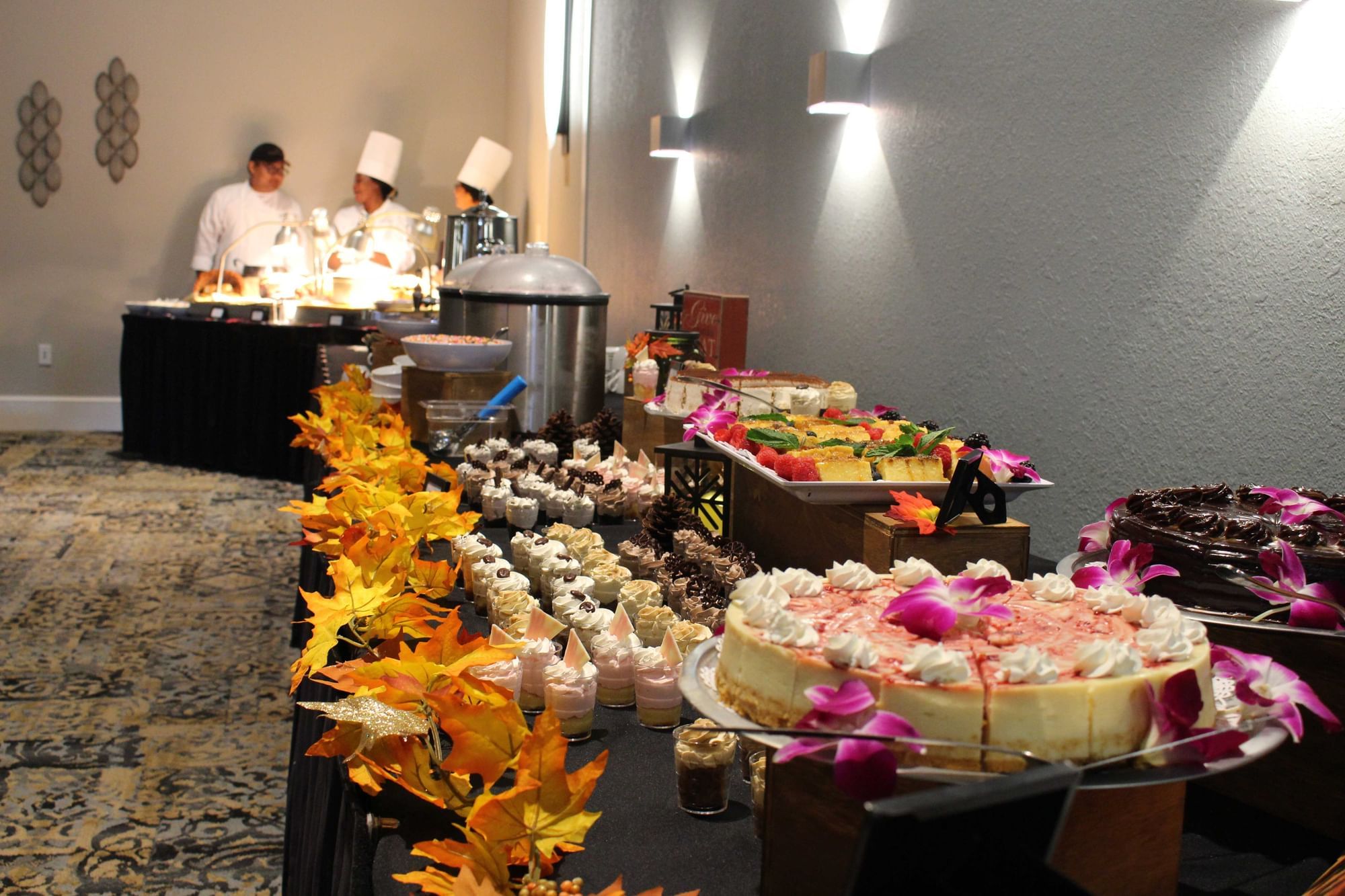 A buffet table with various food items and desserts, including a cake, and two chefs in the background.