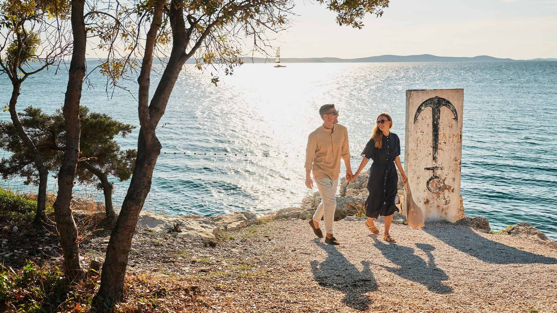 Couple holding hands walking on rocky shore by the sea with a large sign in the background.