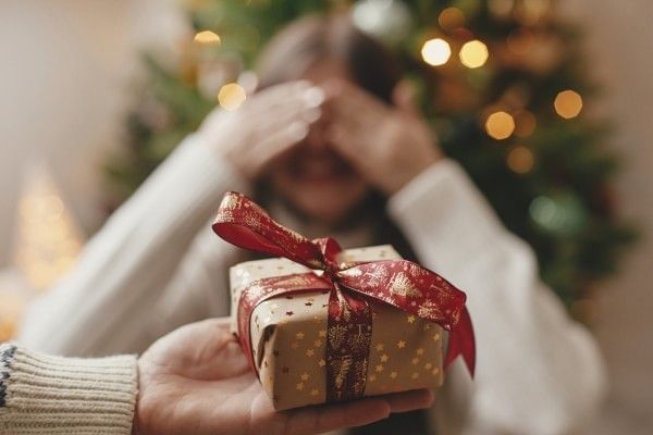 An outstretched hand holds a small wrapped Christmas gift with a red bow in front of a woman covering her eyes with her hands.