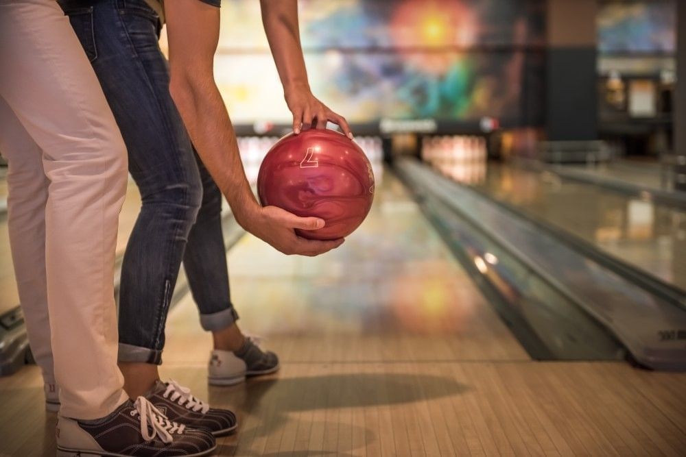 A close-up shot of a man and a woman's legs as they prepare to roll a red bowling ball down a bowling lane together.