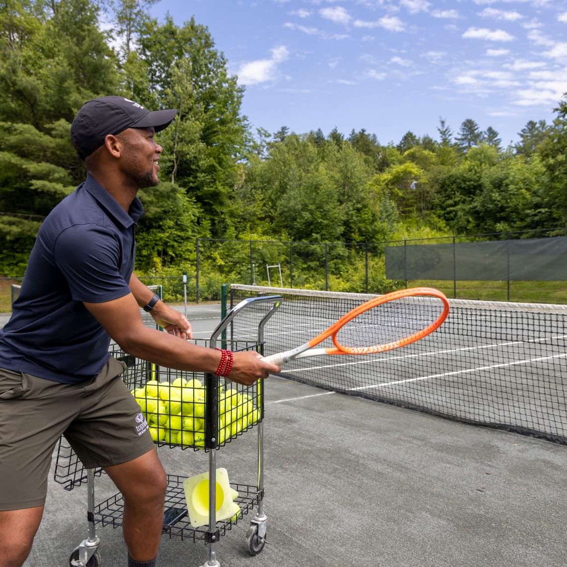 Man holding tennis racket, hitting tennis ball on tennis court with trolley of balls beside him.