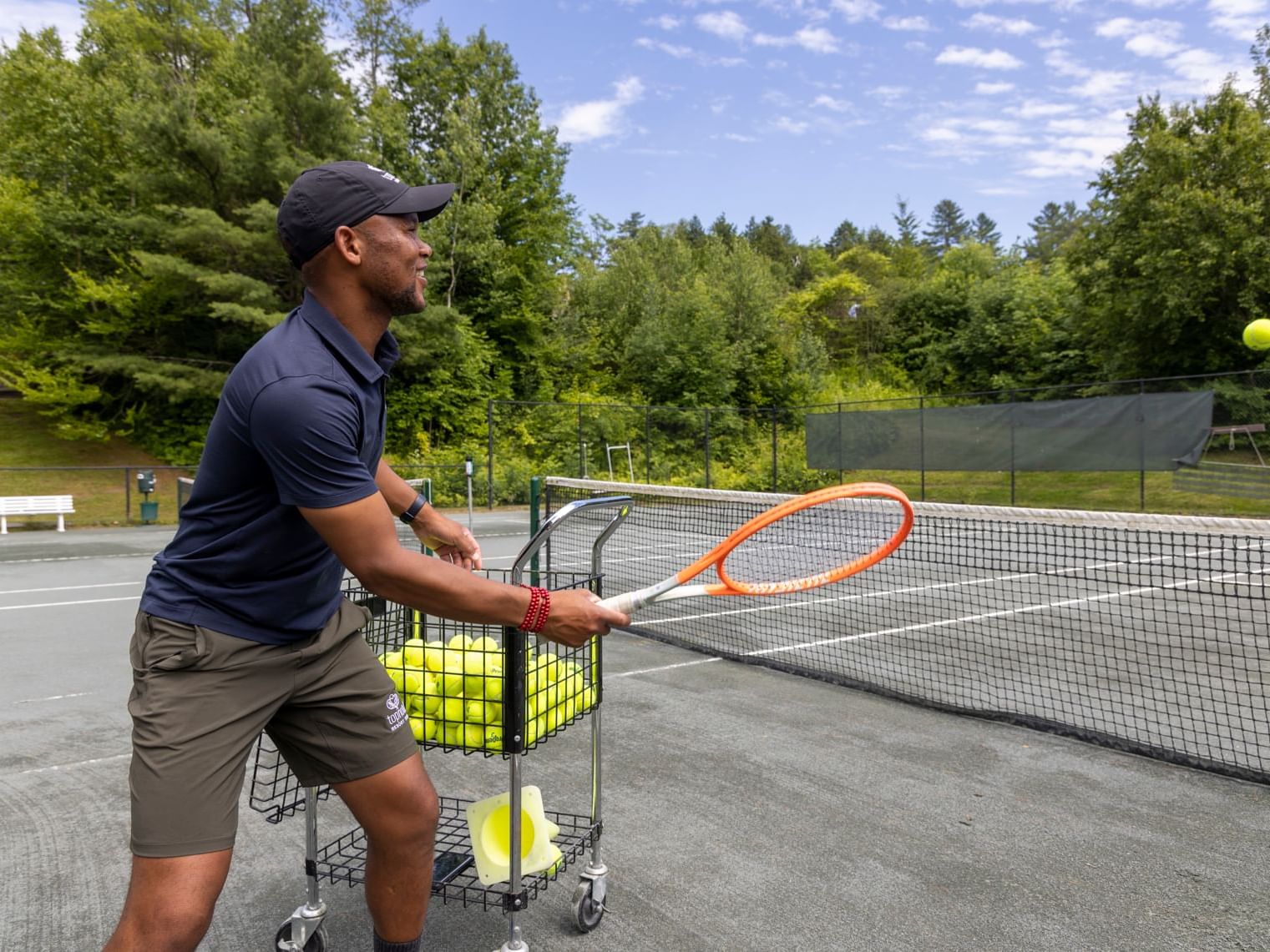 Tennis Director Simba Happy with a tennis ball in the air and cart full of balls.