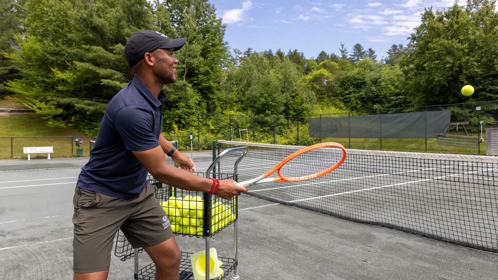 Man holding tennis racket, hitting tennis ball on tennis court with trolley of balls beside him.