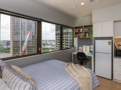 Modern bedroom at UniLodge Brisbane City with bed, desk, fridge, microwave, and city view.