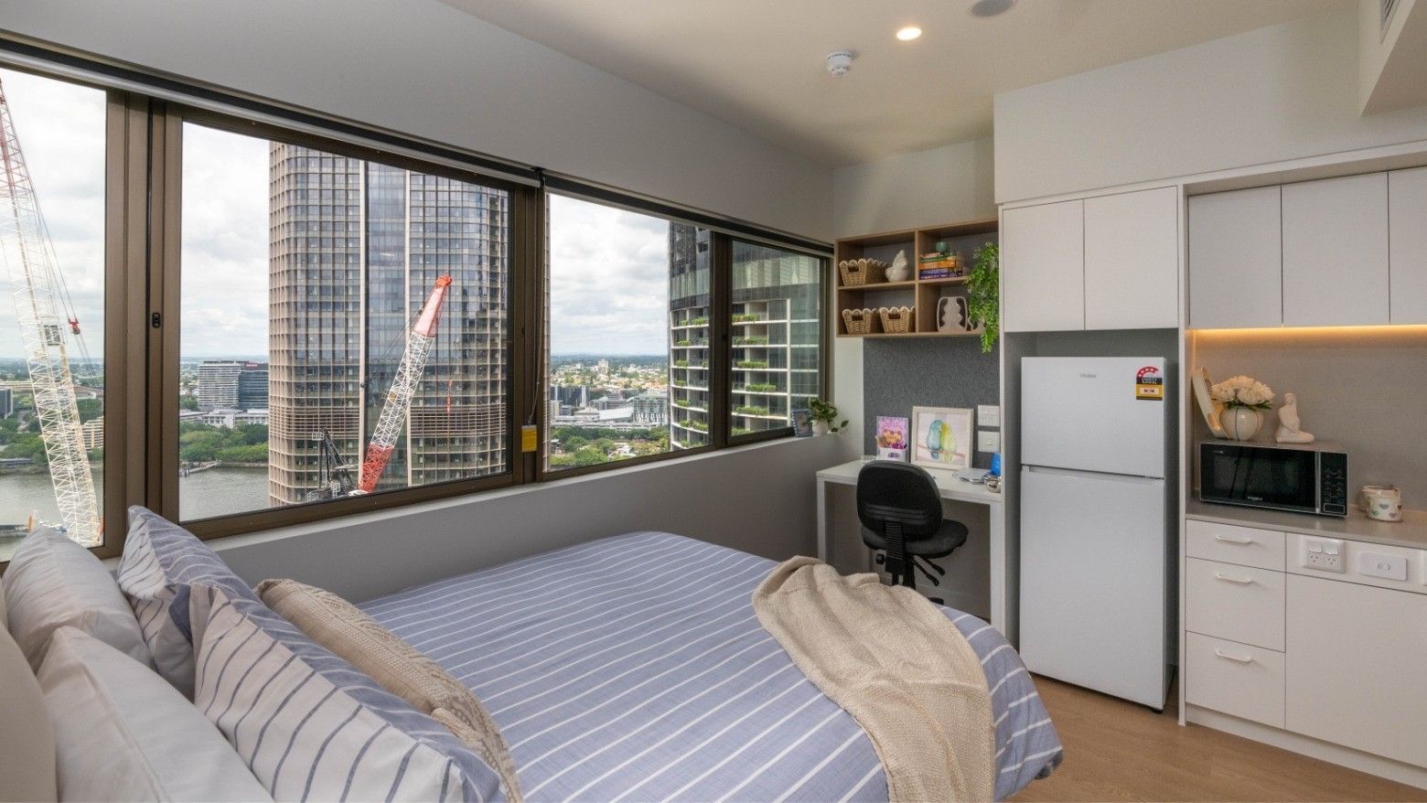 Modern bedroom at UniLodge Brisbane City with bed, desk, fridge, microwave, and city view.