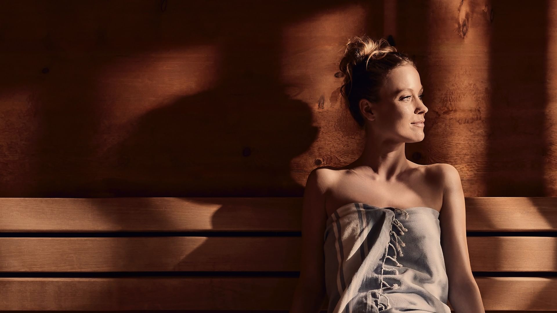 Woman in a blue dress sitting on a wooden bench with sunlight casting shadows on the wall.