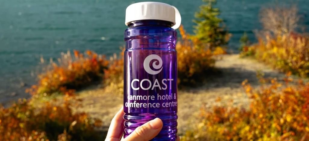 A person holds up a reusable water bottle in front of the mountains on a hiking trail near Canmore.
