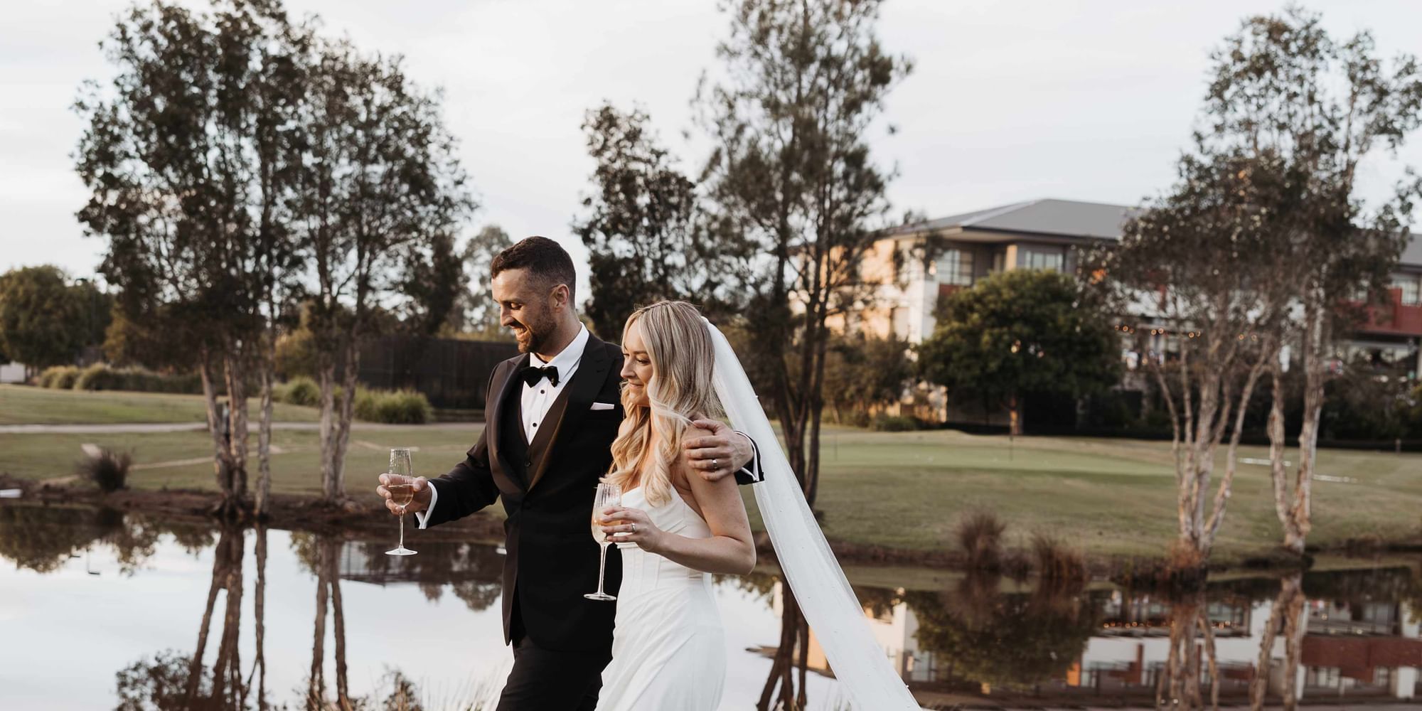 Bride and groom holding champagne glasses by a lake near Mercure Kooindah Waters
