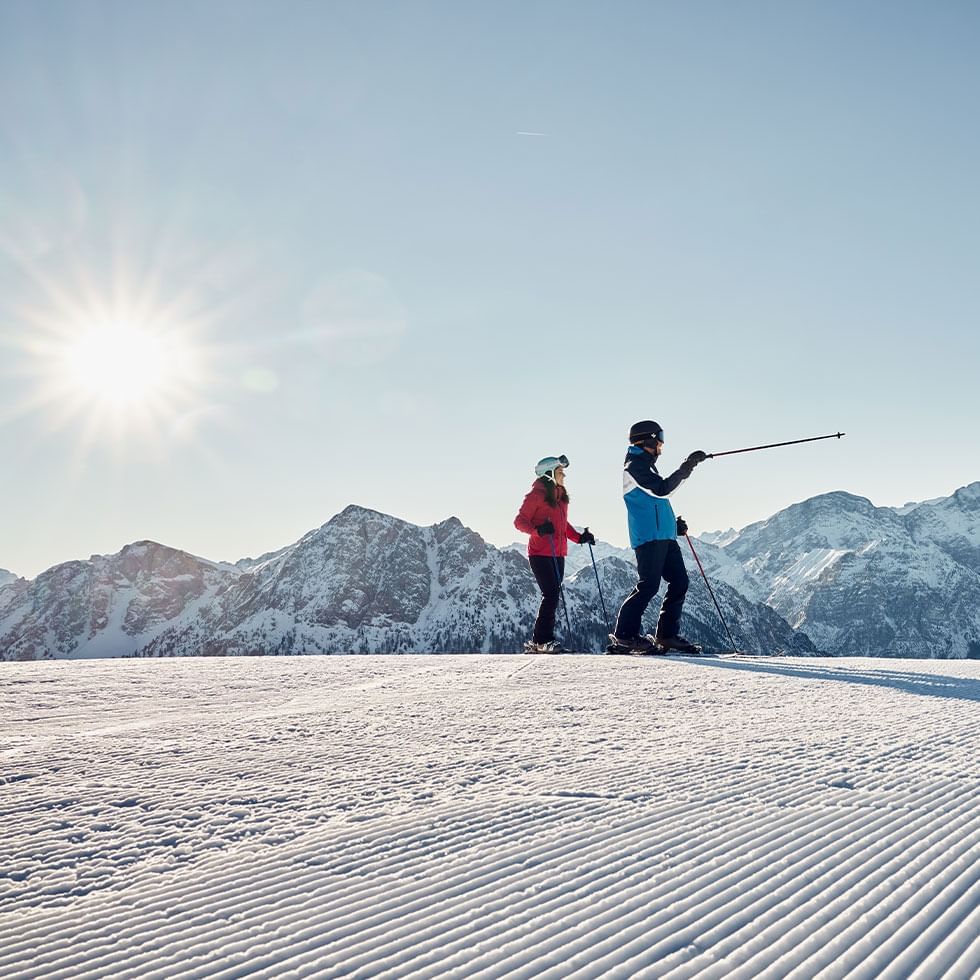Due sciatori su una pista di sci con montagne innevate sullo sfondo sotto il sole.