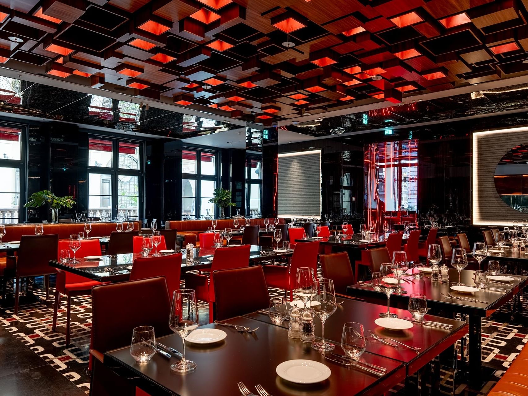 Modern restaurant interior with red chairs, set tables, geometric ceiling panels, and large windows at Titanic Gendarmenmarkt Berlin.