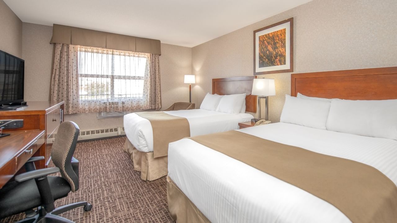 Two beds, desk, chair, window with curtains, TV, and framed artwork in a hotel room.