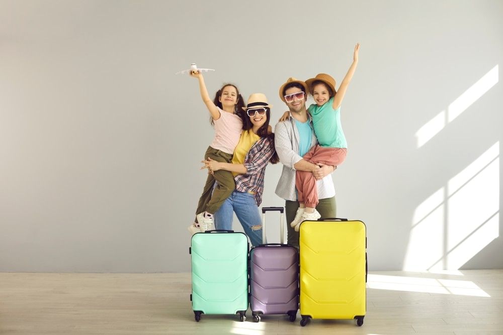 Two parents in sunglasses and hats carry two children, standing in front of three brightly colored suitcases.