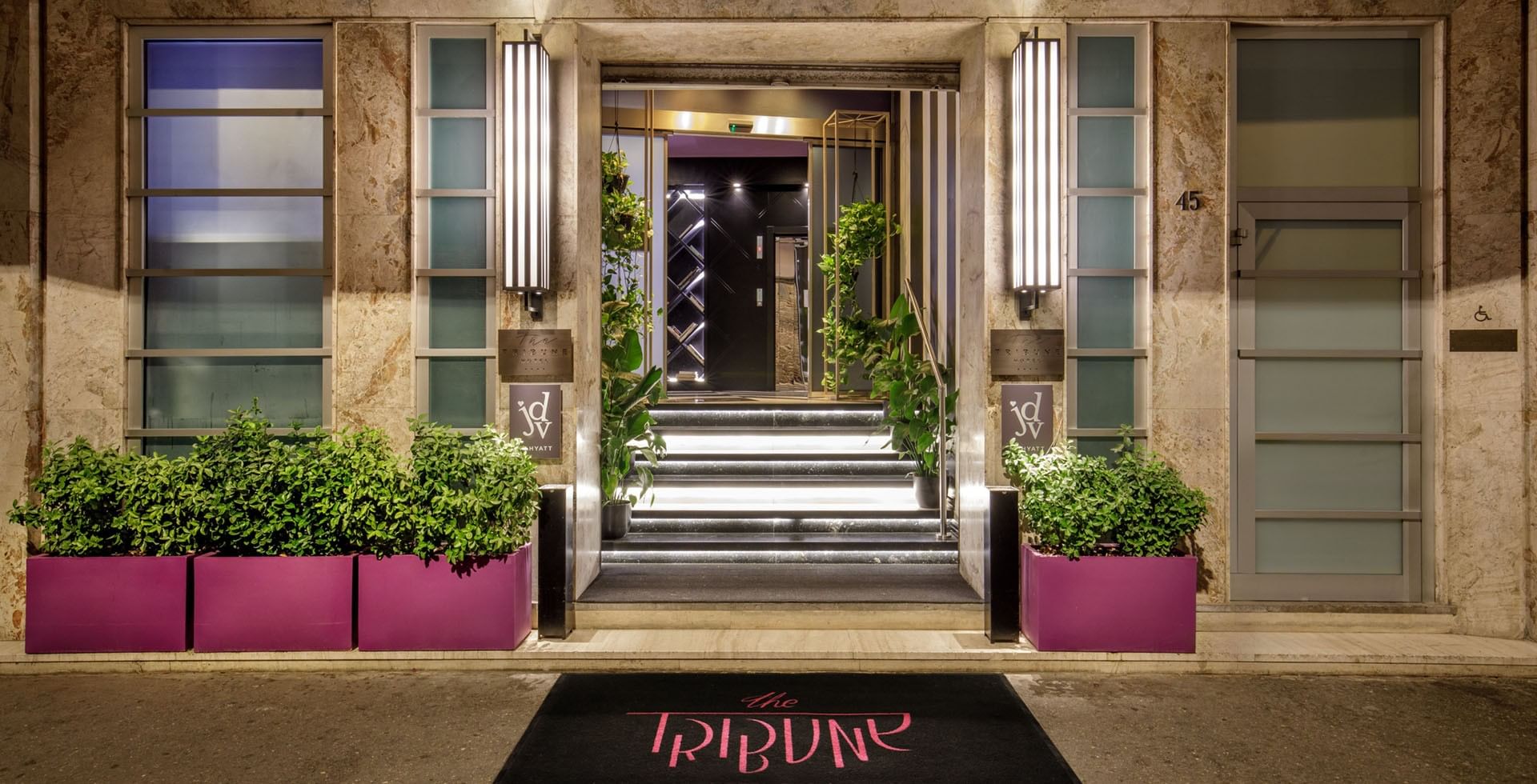 Elegant entrance of Tribune Hotel Rome with illuminated steps, modern décor, and greenery welcoming guests into a stylish contemporary lobby.