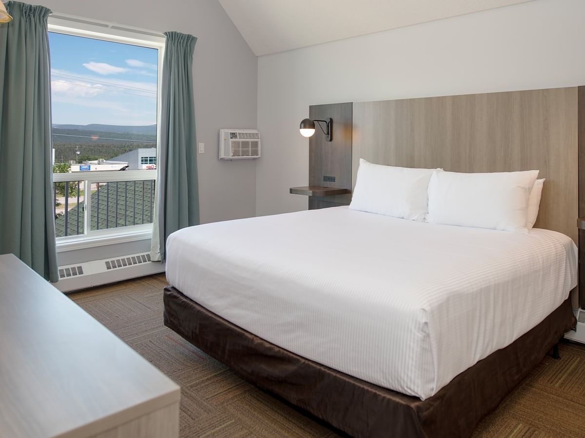 A hotel room with a bed, two pillows, a TV, and a large window with curtains.