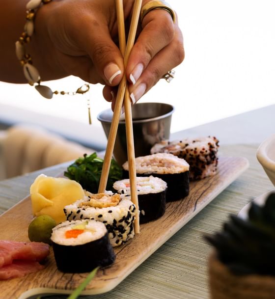 Close-up of a hand using chopsticks to pick up sushi rolls from a wooden platter at Windsong Resort On The Reef