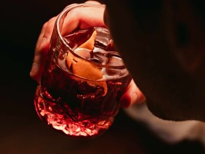 Hand holding a glass of amber-colored cocktail with ice and an orange twist garnish at Amora Herencia Riverwalk Melbourne