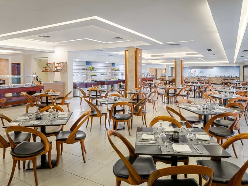 An empty restaurant with numerous tables and chairs, each set with cutlery and glasses, under a modern ceiling design.