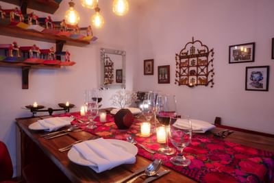 Decorated dining table with red table runner, white plates, cutlery, candles, and wine glasses in a room.
