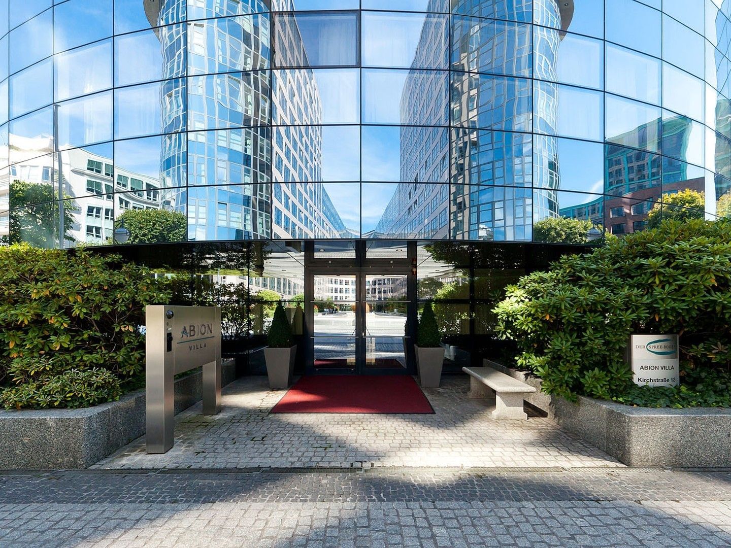 Gläserner Gebäudeeingang mit rotem Teppich, Topfpflanzen und einem Schild mit der Aufschrift 