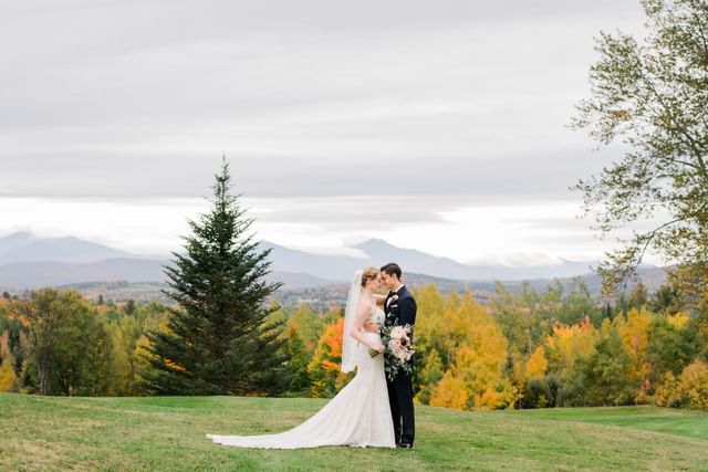 Bride and groom embracing on a grassy hill, near a pine tree and autumn trees at Mountain View Grand Resort & Spa