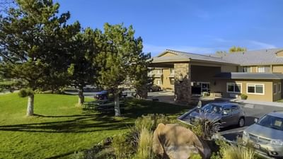 Peaceful Moab Valley Inn entrance with green lawns, shady trees, and parked cars under a bright, clear blue sky