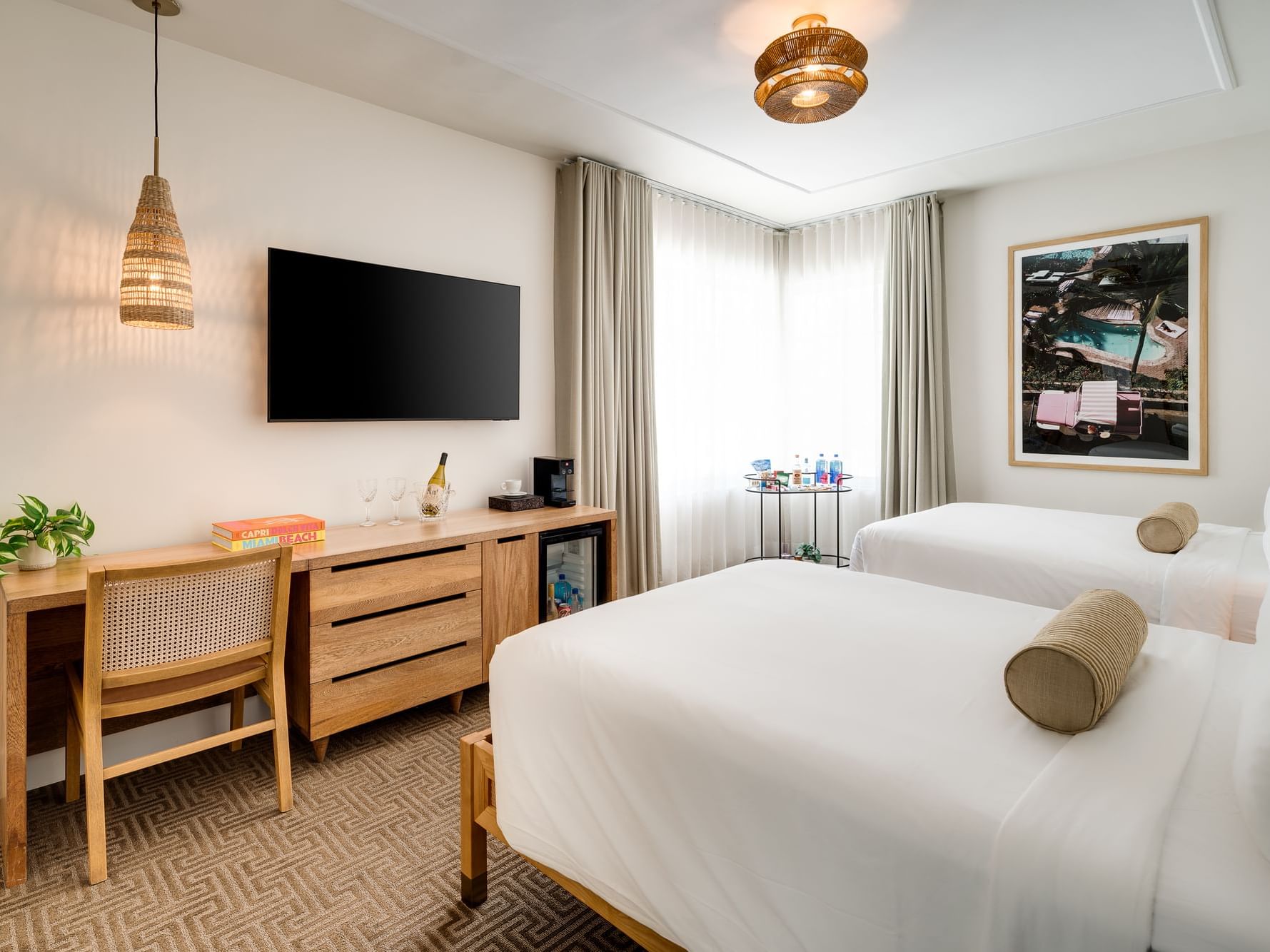 A bright, stylish hotel room with two beds, a desk, chair, TV, and framed artwork on the wall.