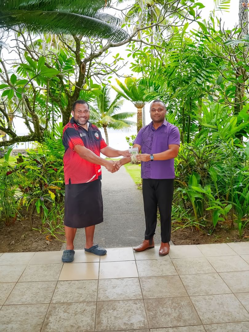 Two men shaking hands and exchanging an envelope on a tiled patio surrounded by lush tropical greenery.