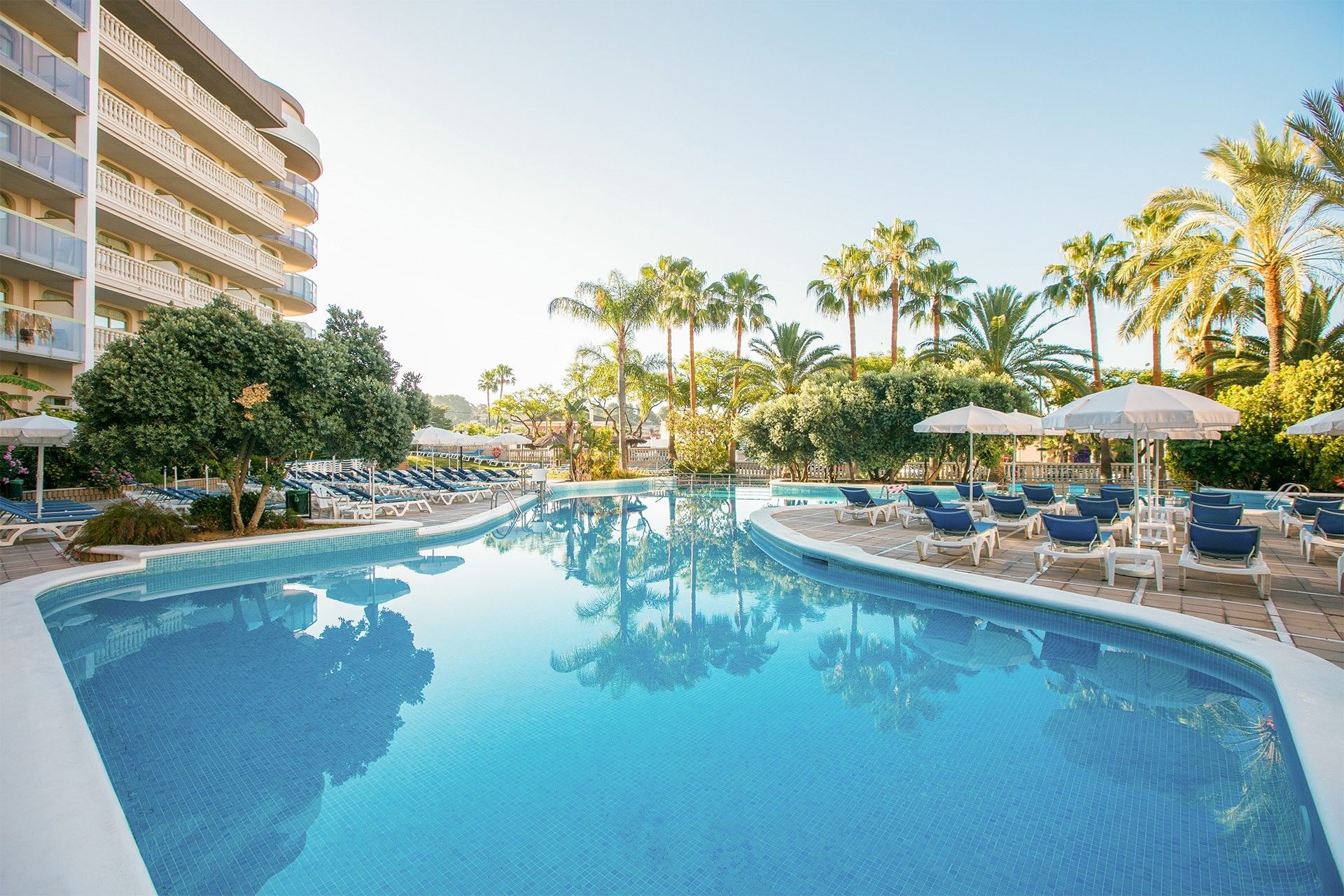 A bright, sunlit hotel pool area with turquoise water, palm trees, white umbrellas, and a multi-story building.