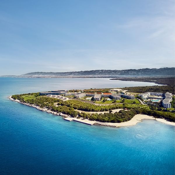 Aerial view of a coastal resort with buildings and greenery surrounded by blue sea at Falkensteiner Group