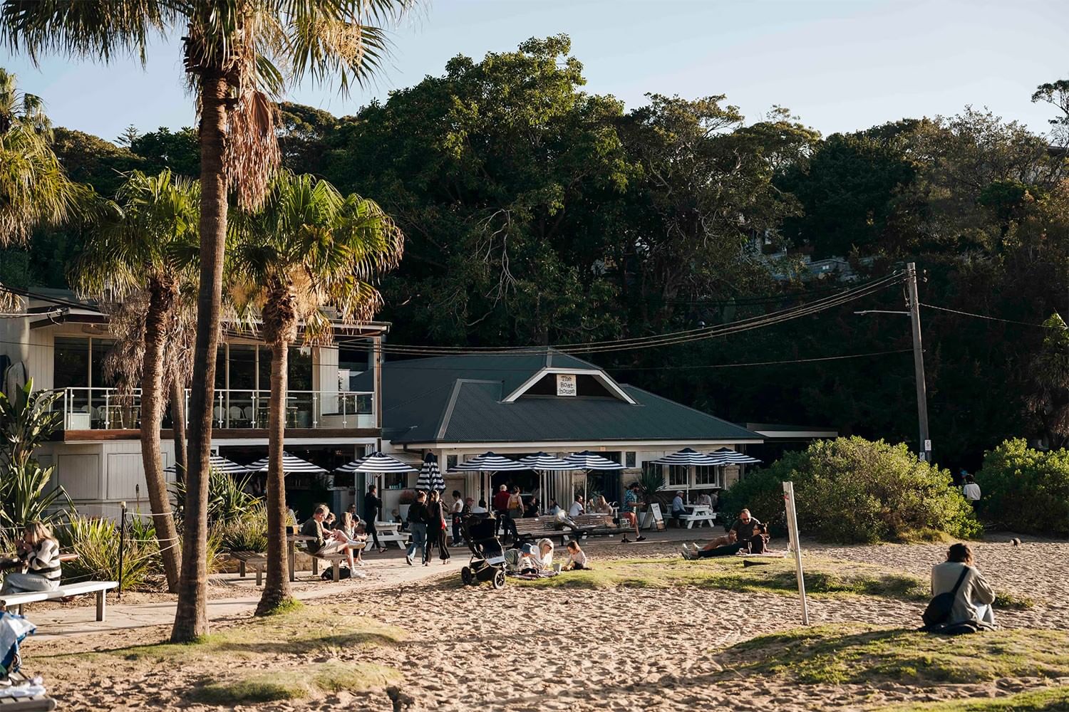 The Boathouse at Elysium Hotels is beside a sandy beach with palm trees and outdoor seating under striped umbrellas